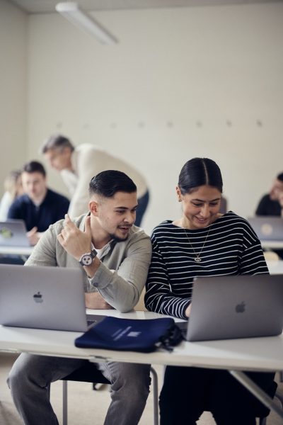 Två elever sitter med sina datorer i ett klassrum