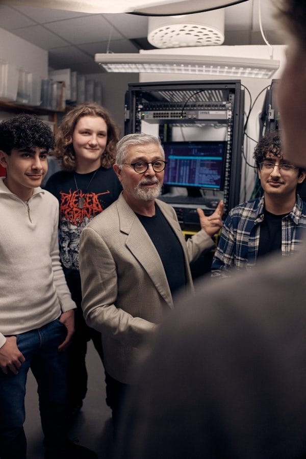 Tre elever och en lärare står i ett rum med en datorskärm bakom sig