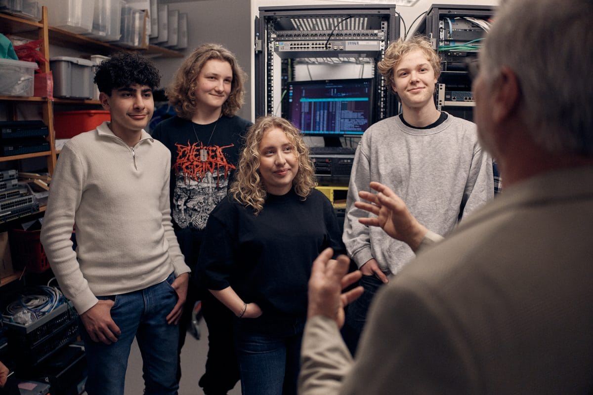 Fyra elever lyssnar på en lärare i ett rum med datorer och sladdar